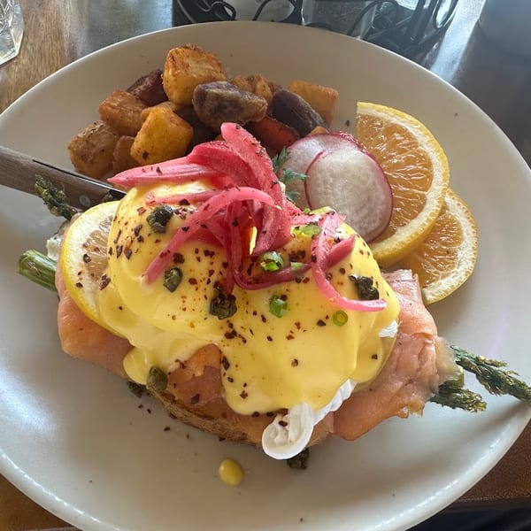 Eggs Benedict with salmon, asparagus, home fries, orange slices, and radishes on a white plate.