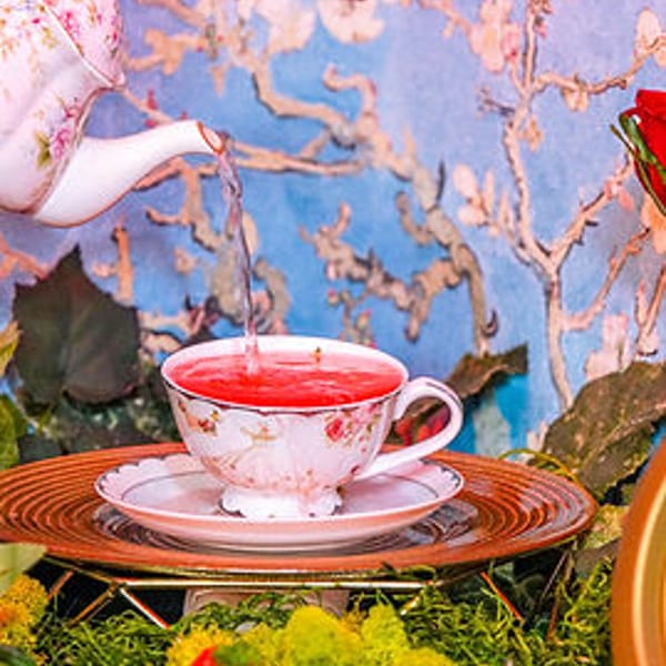 Teapot pouring red tea into a floral cup, surrounded by pastries and moss against a floral backdrop.