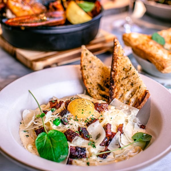 Creamy pasta with bacon, peas, parmesan, raw egg yolk, and toasted bread, with a shrimp dish in the background.