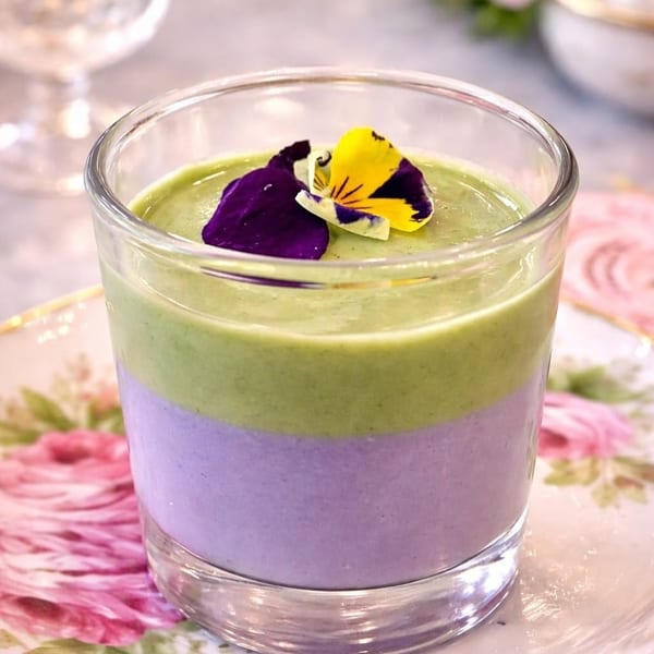 A vibrant purple and green layered dessert in a glass, garnished with edible pansy flowers.