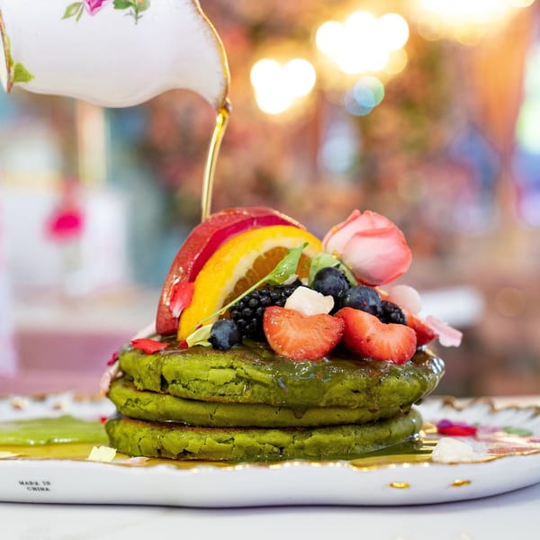 Syrup pouring from a floral teapot onto green pancakes topped with fresh fruit and a pink rose.