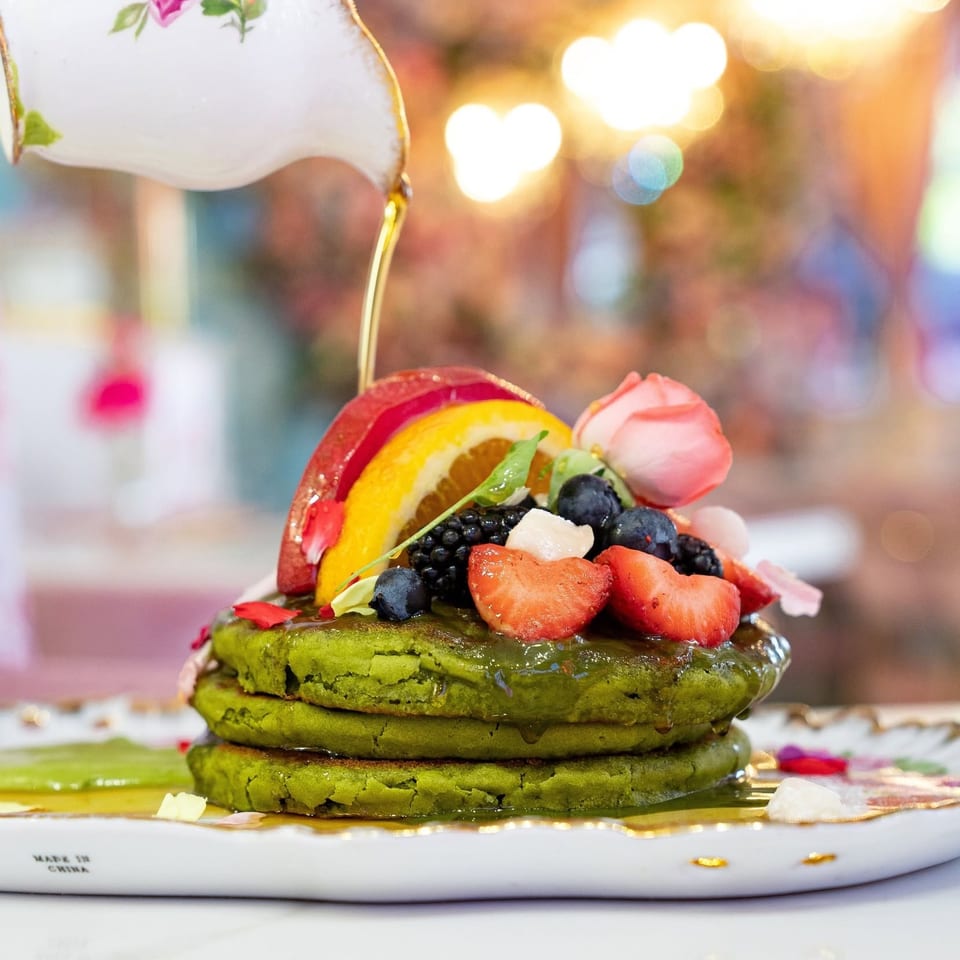 Syrup pouring from a floral teapot onto green pancakes topped with fresh fruit and a pink rose.