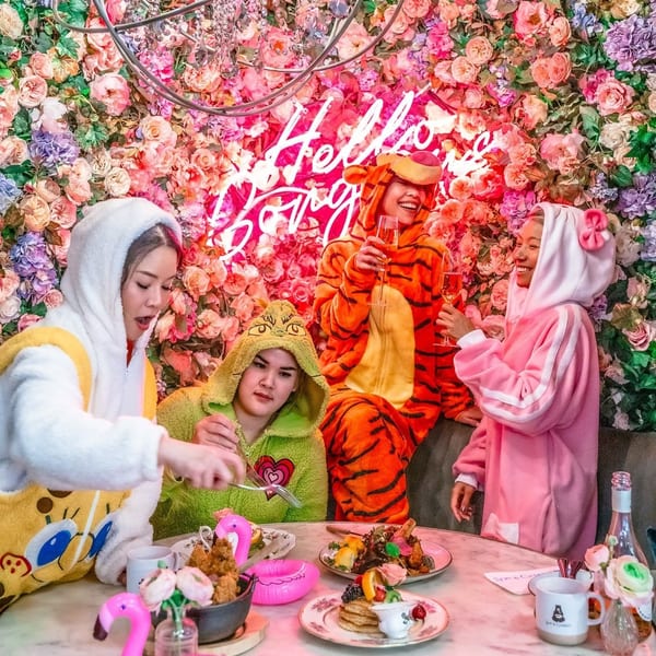Four friends in animal onesies enjoy brunch and champagne against a vibrant floral wall with a neon sign.