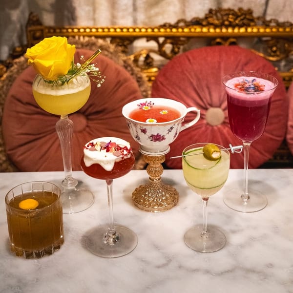 Five colorful cocktails, including one with a yellow rose, on a marble table with ornate gold and red seating.
