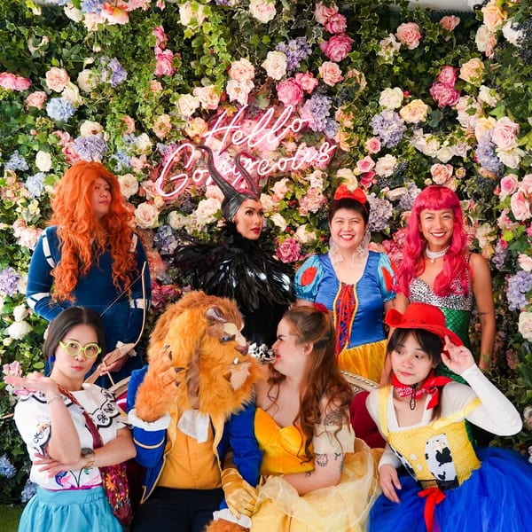 Seven people in Disney character costumes pose in front of a colorful floral wall with a neon sign.